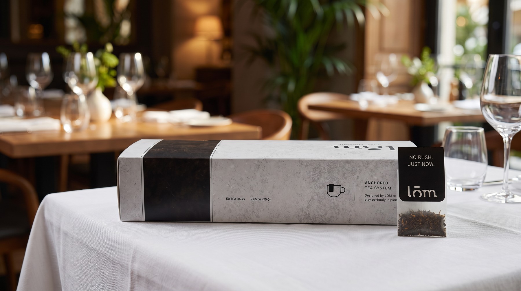 Caja de té LŌM y sobre individual sobre mesa de mantel blanco en un restaurante, con copas de vino de fondo